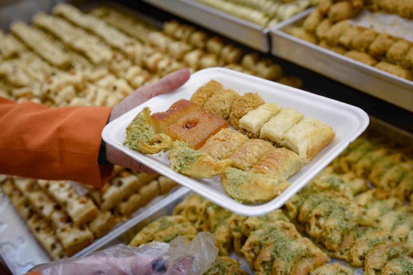 A hand holding a white tray of assorted baklava and semolina cake over the pyramid displays at Abd al-Hadi Sweets